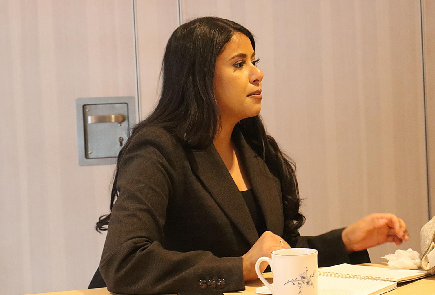 En kvinna med långt mörkt hår i svart kavaj sitter vid ett bord med en anteckningsbok och en kaffekopp och tittar åt sidan.