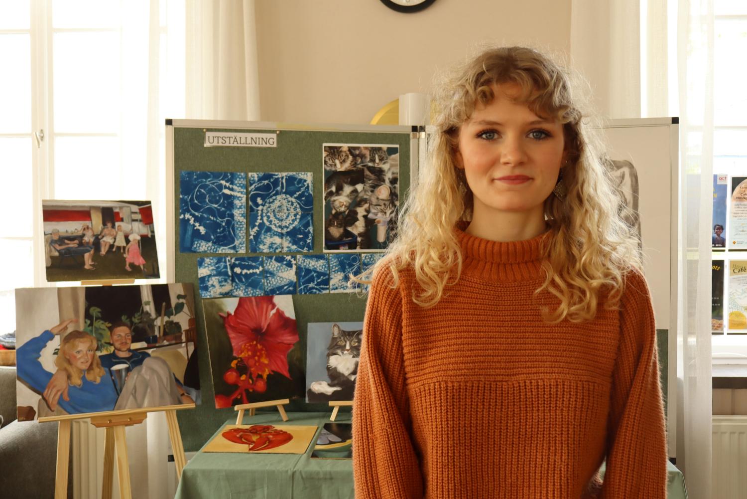 En kvinna i en orange tröja står inomhus framför en konstutställning med målningar, foton och ritningar på stafflier och en tavla märkt 
