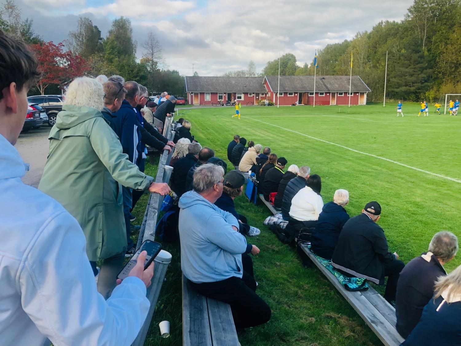 Åskådare sitter och står på träbänkar och tittar på en fotbollsmatch på en gräsplan nära en röd byggnad och omgiven av träd.