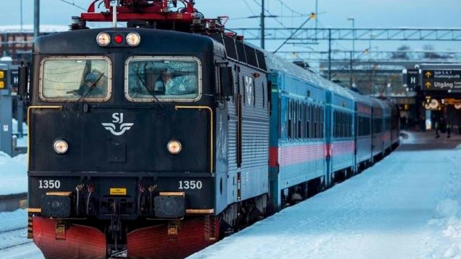 Ett svart SJ-tåg med numret 1350 står stilla på en snötäckt perrong, med kontaktledningar och skyltar synliga i bakgrunden.