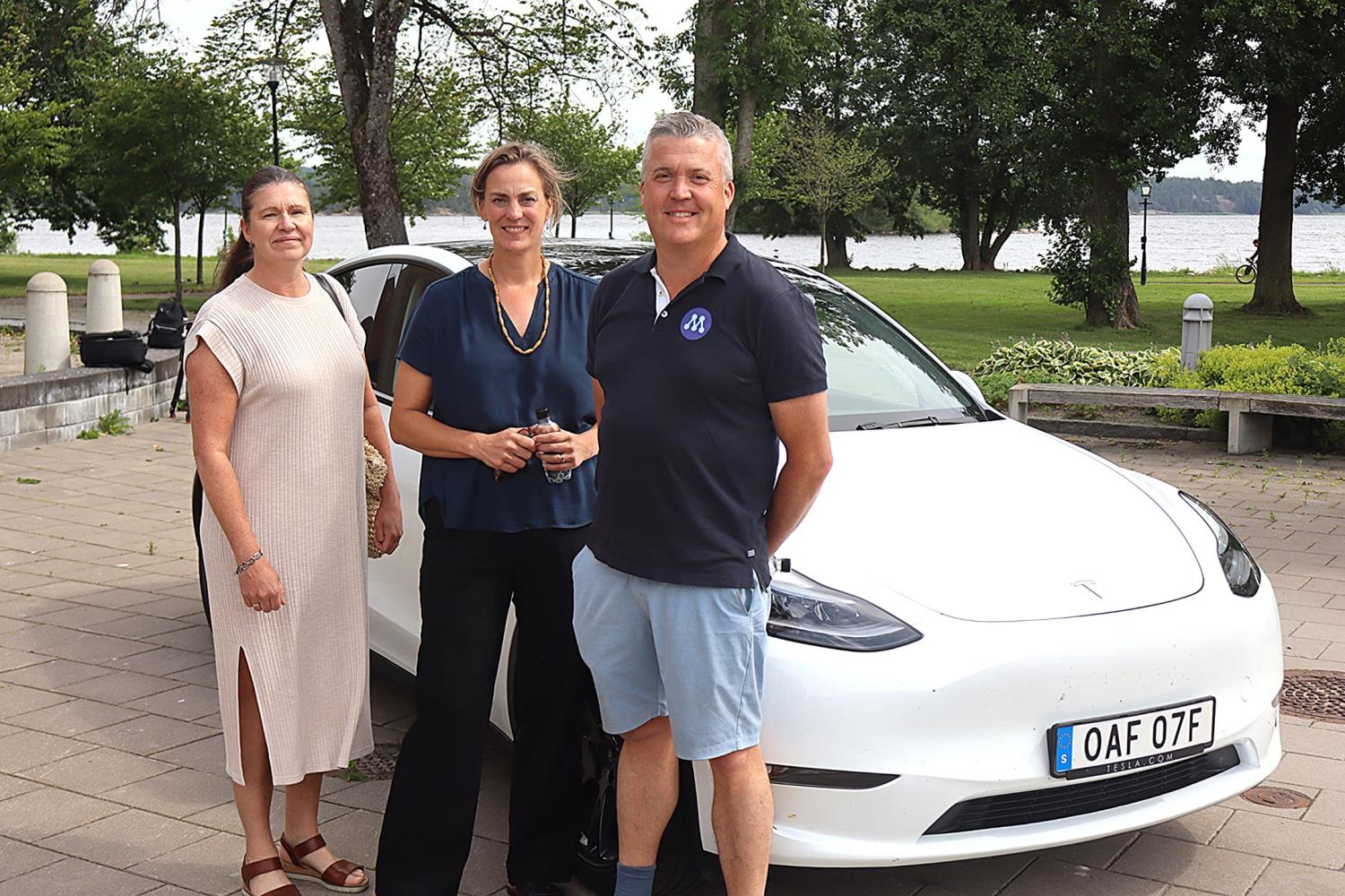 Tre vuxna står framför en vit Tesla-bil som står parkerad nära en park med träd och en sjö i bakgrunden.