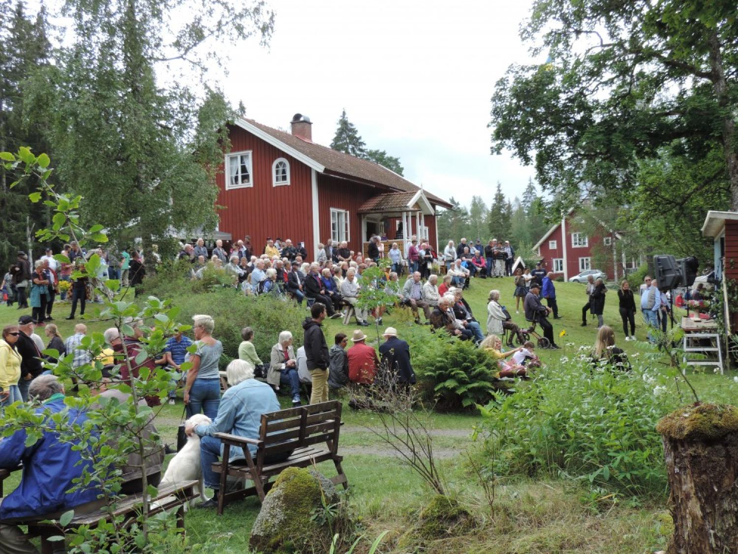 En samling människor, inklusive familjer och individer, sittande på bänkar och gräs utanför röda trähus i en lantlig miljö, omgiven av grönska och träd.