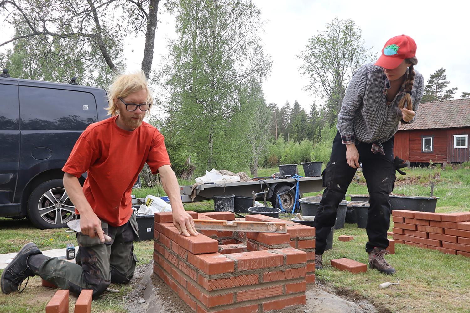 Två personer bygger en liten tegelkonstruktion utomhus; en placerar ut tegelstenar medan den andra står i närheten och tittar på. Byggverktyg och material ligger utspridda runt omkring.