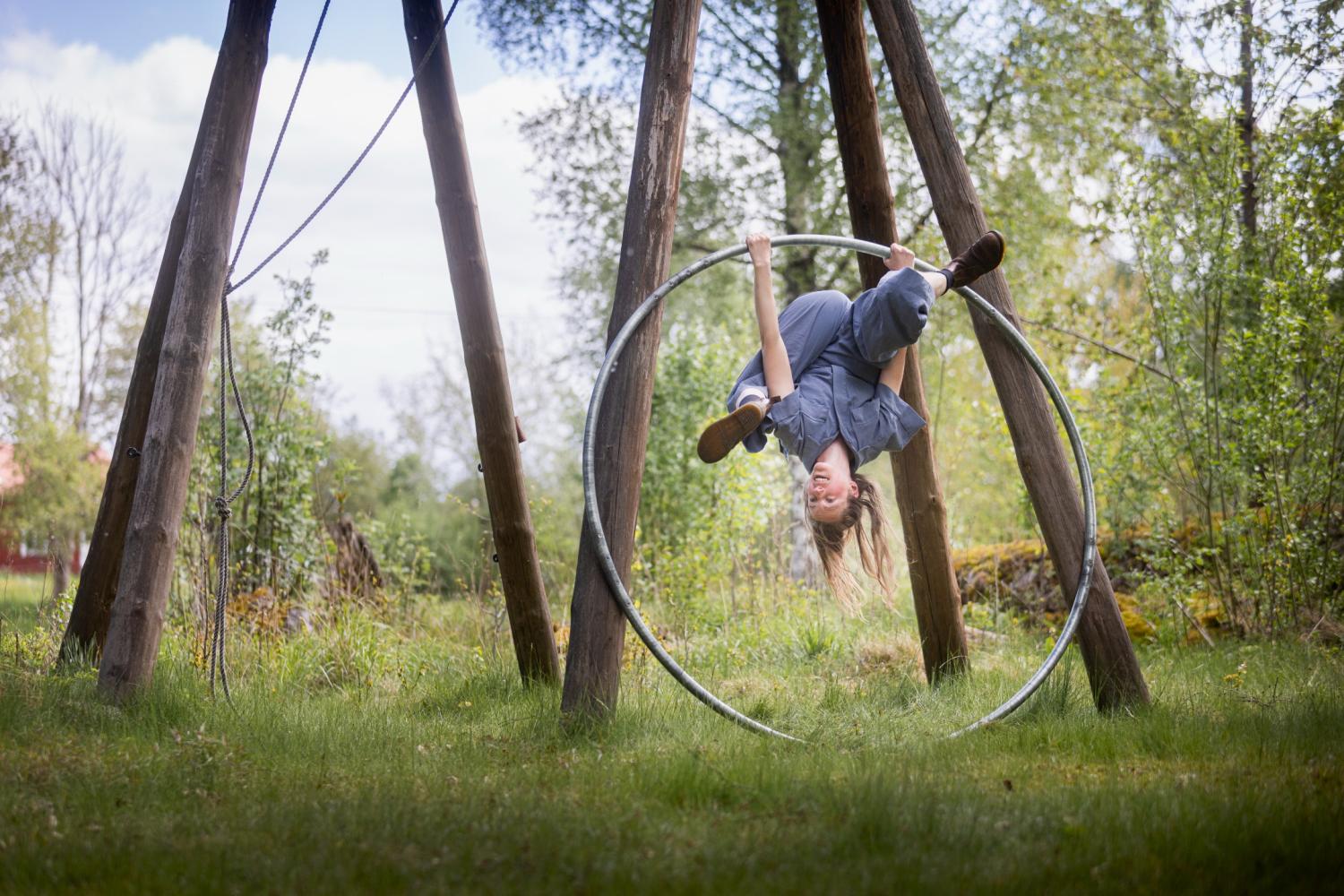 En gråklädd person hänger upp och ner i en metallring utomhus, omgiven av gräs och träd.