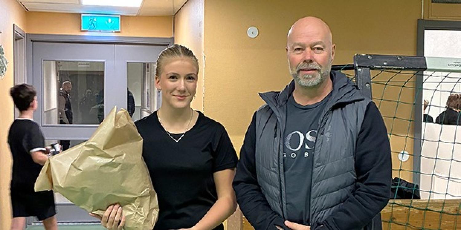 En ung kvinna med en bukett i handen står bredvid en äldre man i en korridor på ett gym, med människor och sportutrustning i bakgrunden.