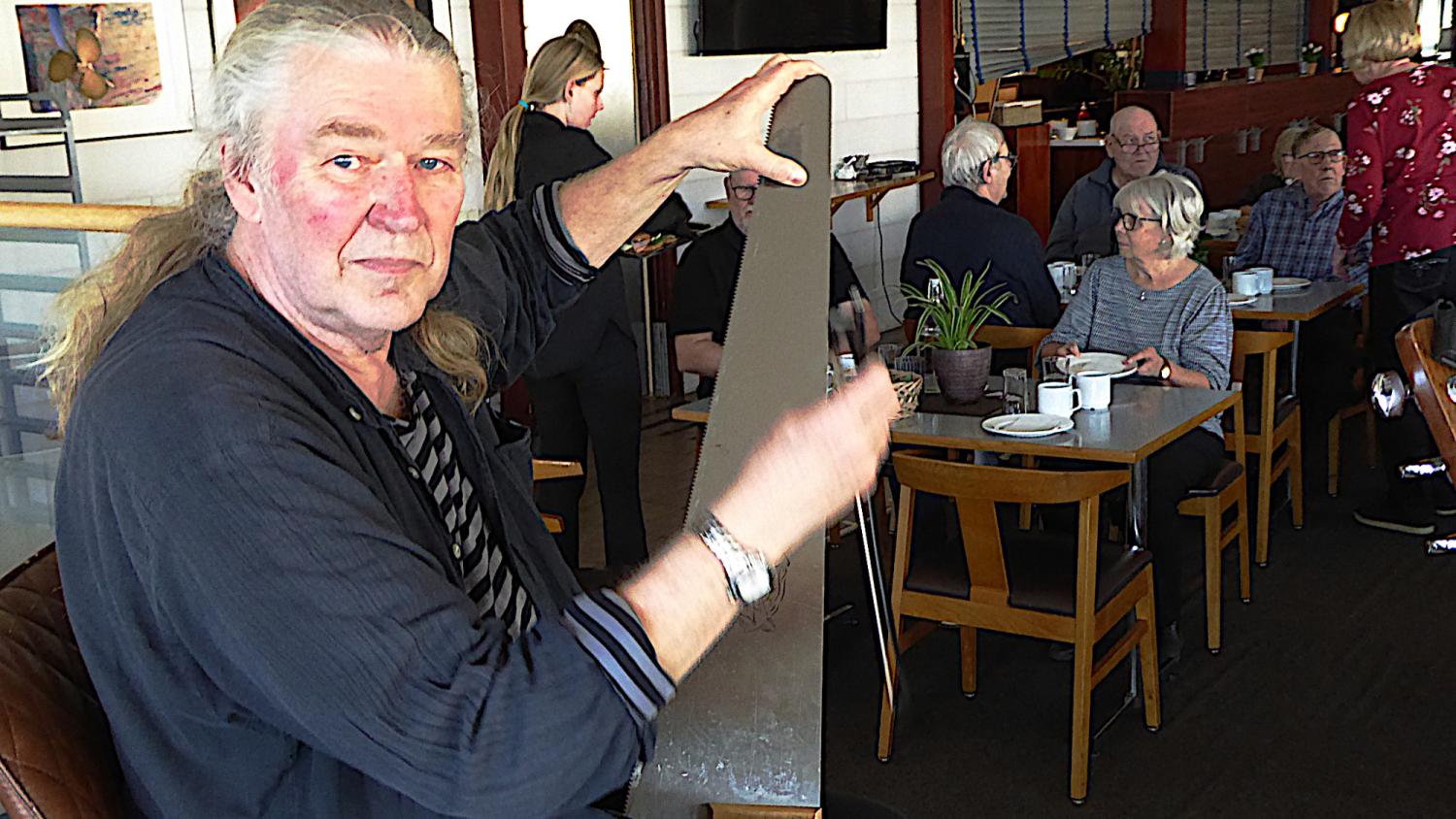 En man sitter på ett café och håller i och böjer en såg, medan andra gäster sitter och samtalar vid bord i bakgrunden.