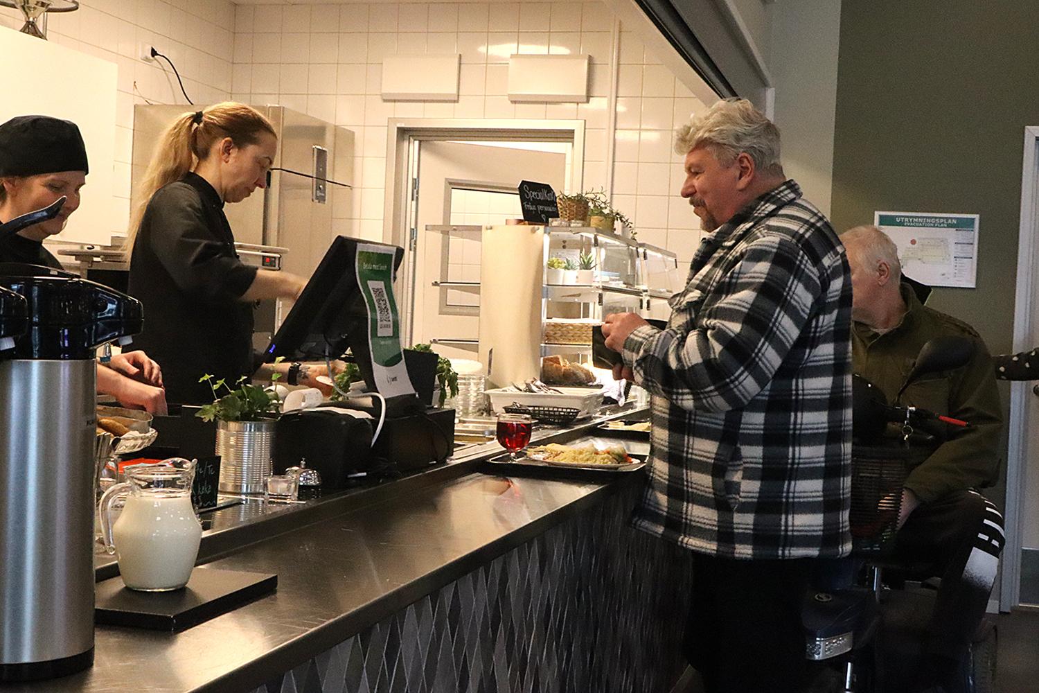 En man i rutig jacka står vid en kafédisk med mat på en bricka medan en anställd serverar honom. En annan man sitter i bakgrunden.