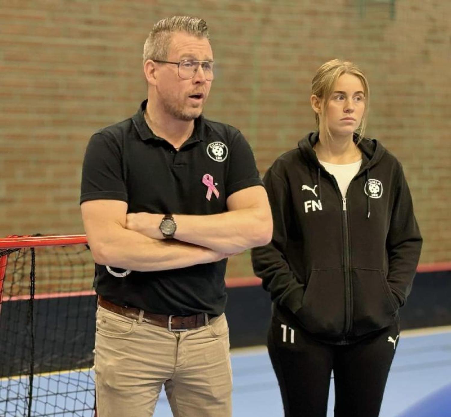 En man och en ung kvinna står inomhus vid ett sportnät; mannen pratar med armarna i kors och kvinnan bär en träningsoverall med händerna i fickorna.