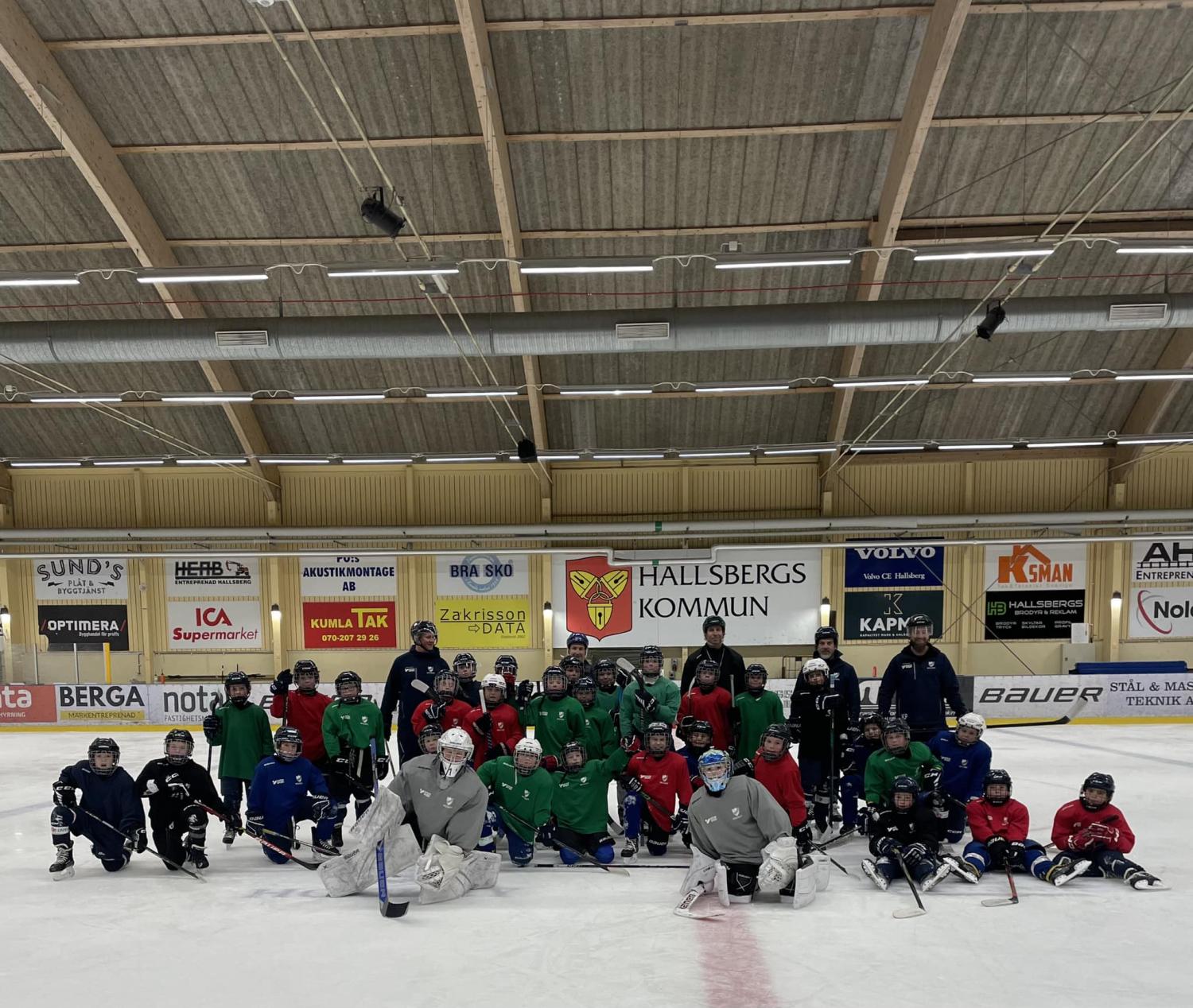 En grupp människor, klädda i färgglada hockeykläder, poserar på en ishall inomhus. Banderoller och annonser syns på väggarna bakom dem.