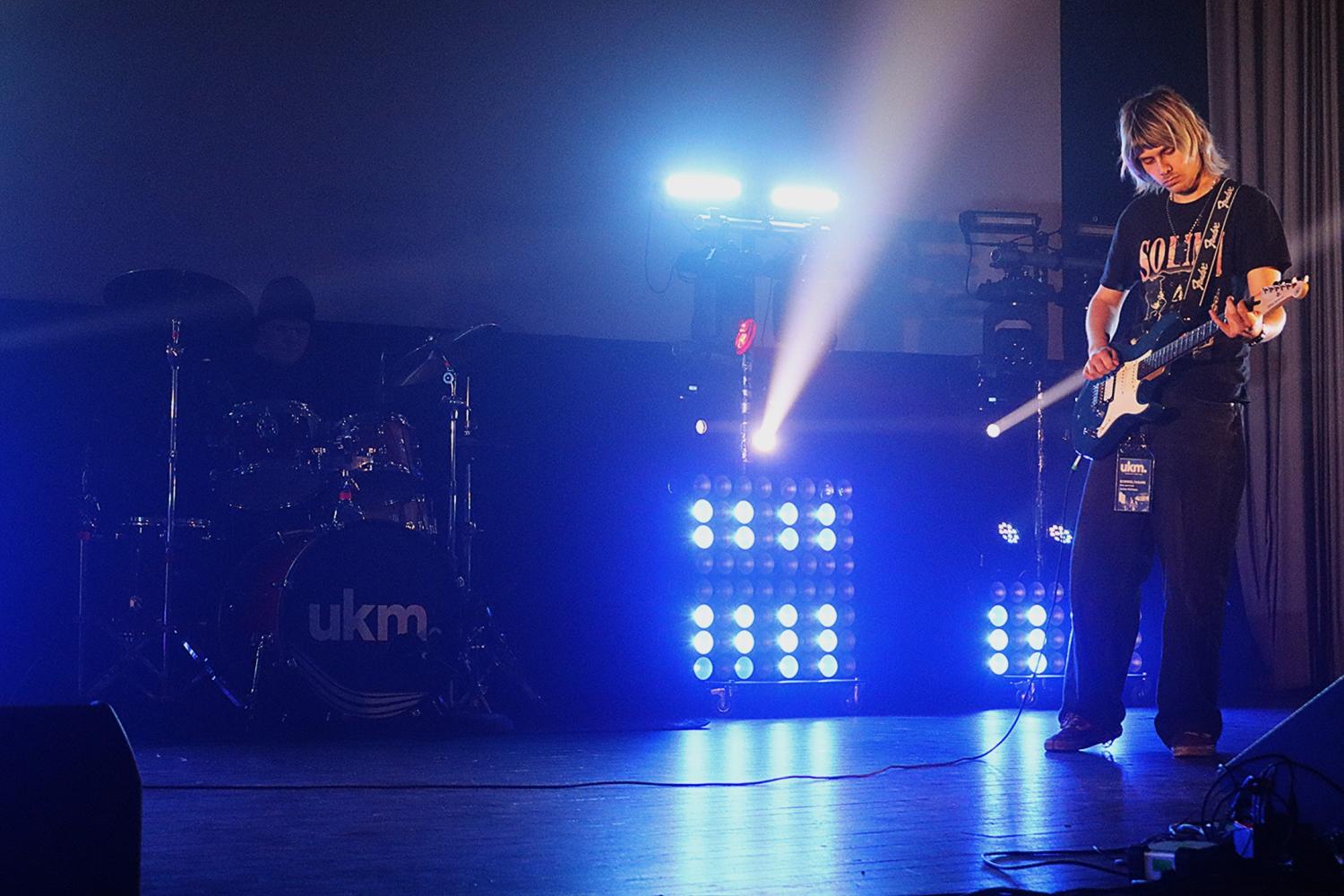 En gitarrist spelar elgitarr på en scen med blå belysning, medan en trummis är delvis synlig i bakgrunden.