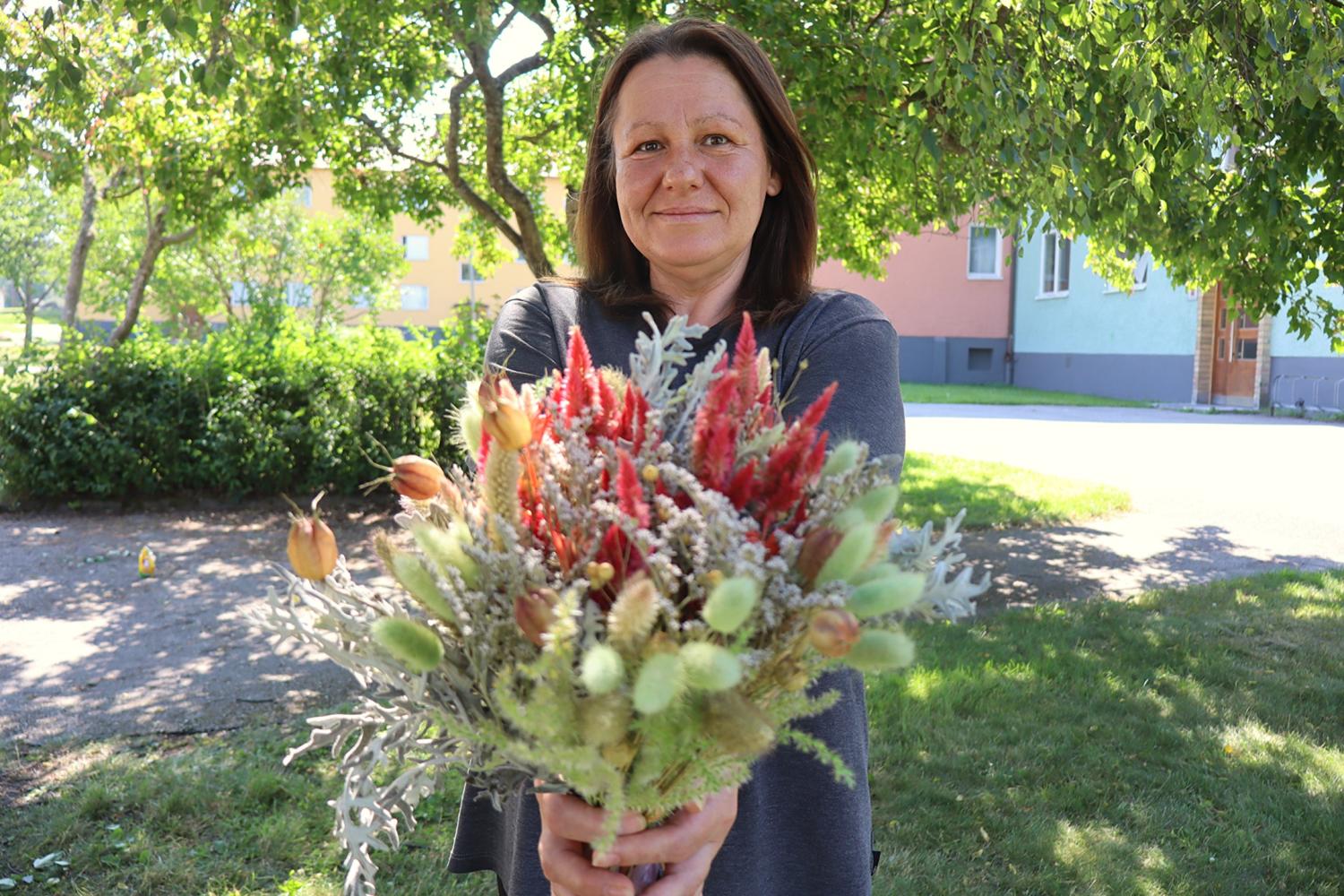 En kvinna står utomhus med en bukett bestående av olika blommor och växter i händerna, medan träd och byggnader syns i bakgrunden.