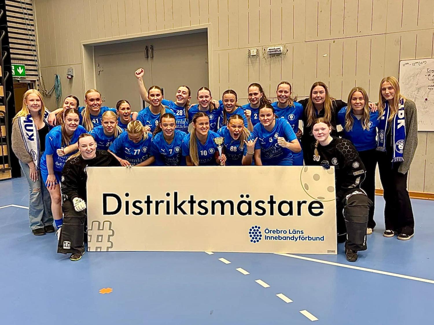 Ett damidrottslag i blå uniformer poserar inomhus bakom en stor skylt med texten 