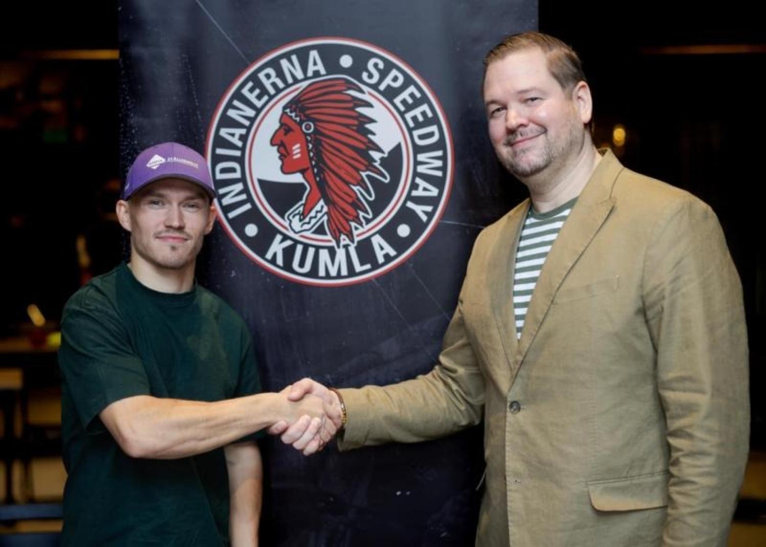 Två män skakar hand framför en Indianerna Speedway Kumla-logotyp, den ene i lila keps och vardagskläder, den andre i beige kavaj och randig skjorta.
