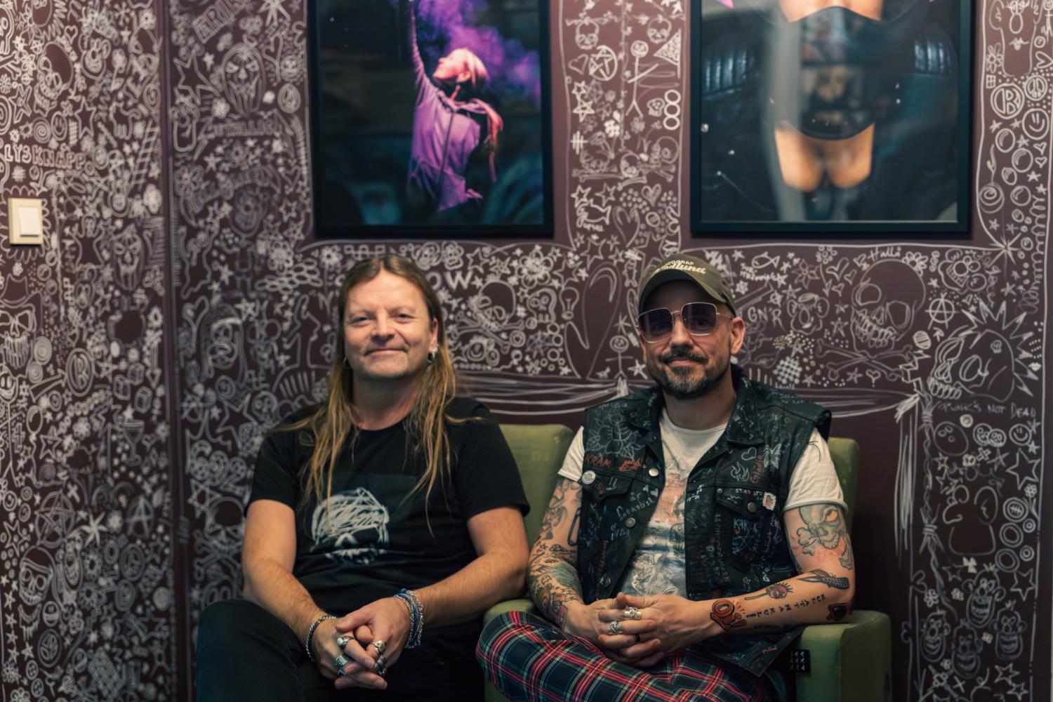 Två män med tatueringar och långt hår sitter på gröna stolar framför en vägg täckt av klotter, med två inramade foton hängande ovanför dem.