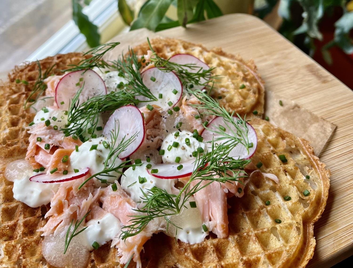 Närbild på en våffla toppad med strimlad lax, skivade rädisor, gräslök, dill och en klick krämig sås, serverad på en träbräda.