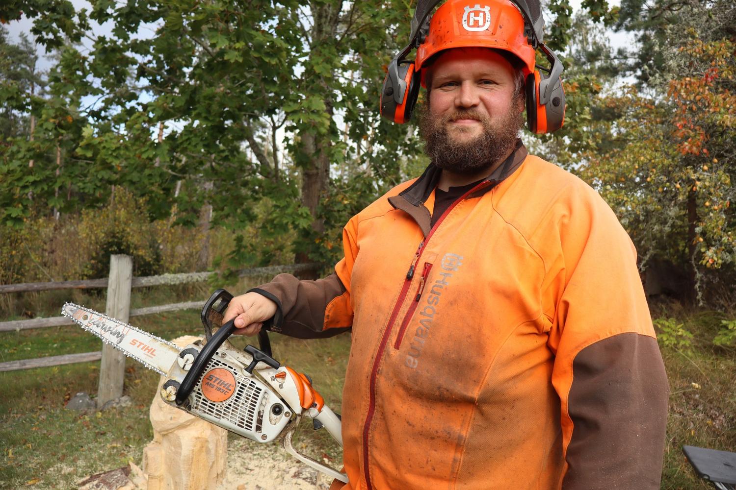 En person i orange skyddsutrustning står utomhus med en motorsåg i handen, medan träd, ett staket och sågspån syns i bakgrunden.