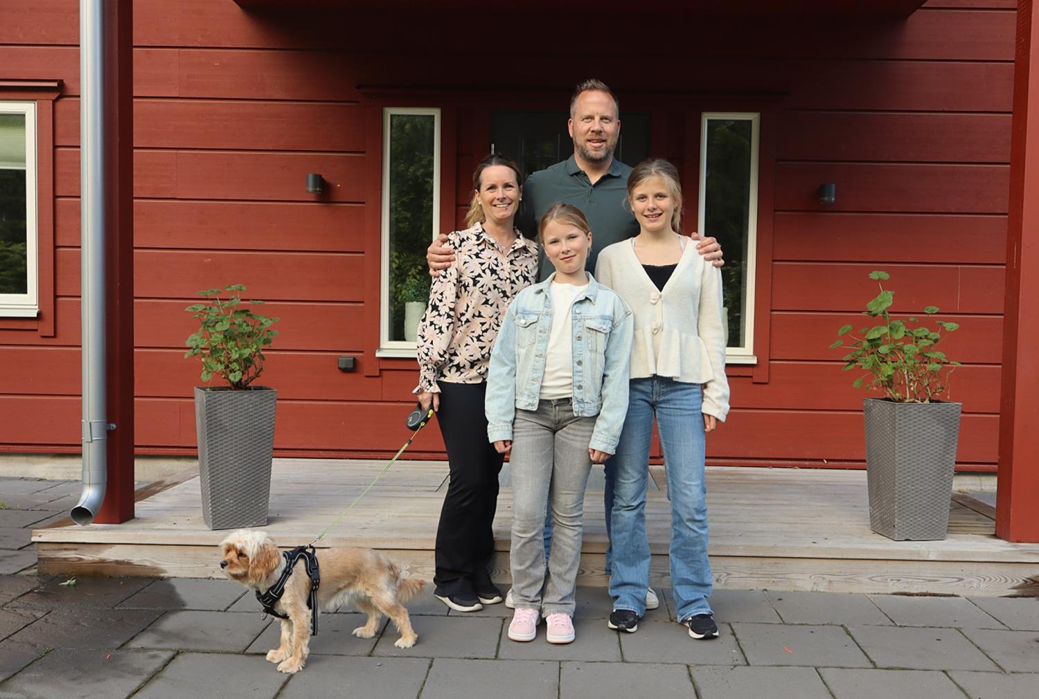 En familj på fyra personer står framför ett rött hus, med två krukväxter och en liten hund i koppel till vänster.