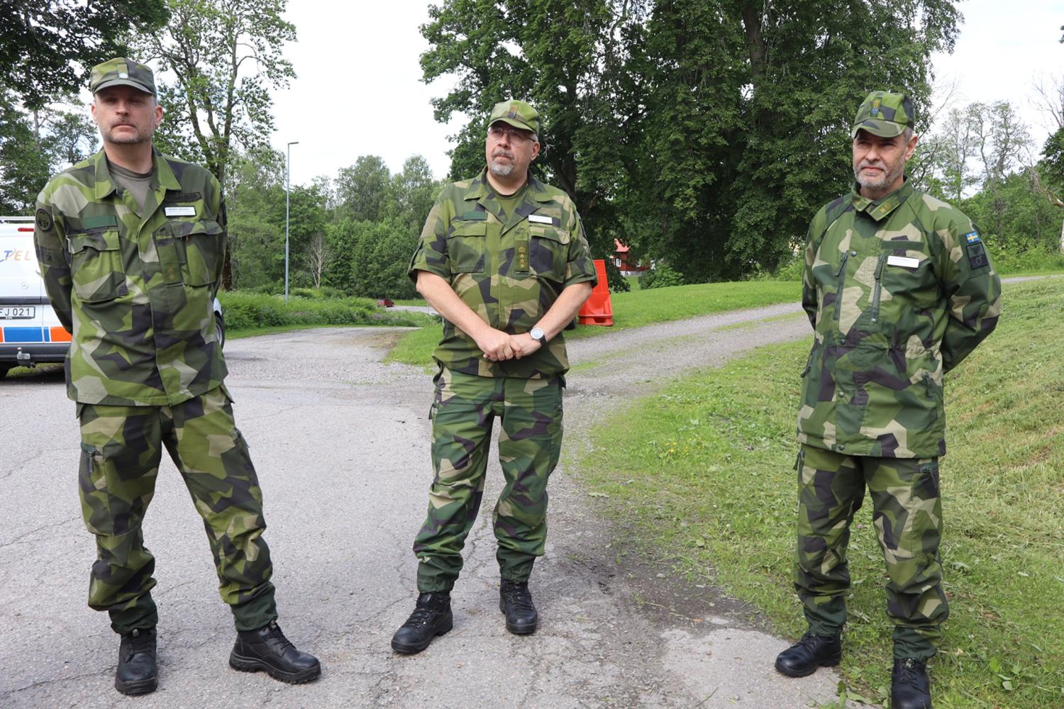 Tre män i kamouflageuniformer står på en asfalterad gångväg utomhus, med träd och gräs i bakgrunden.