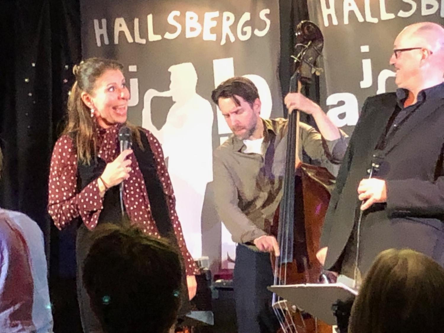 En kvinna med mikrofon står tillsammans med två män, varav en spelar kontrabas, på scenen framför 
