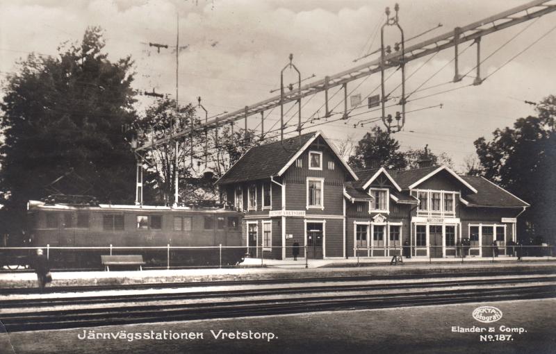 Svartvitt foto av Vretstorps järnvägsstation med ett tåg till vänster, omgivet av träd och elektriska kontaktledningar.