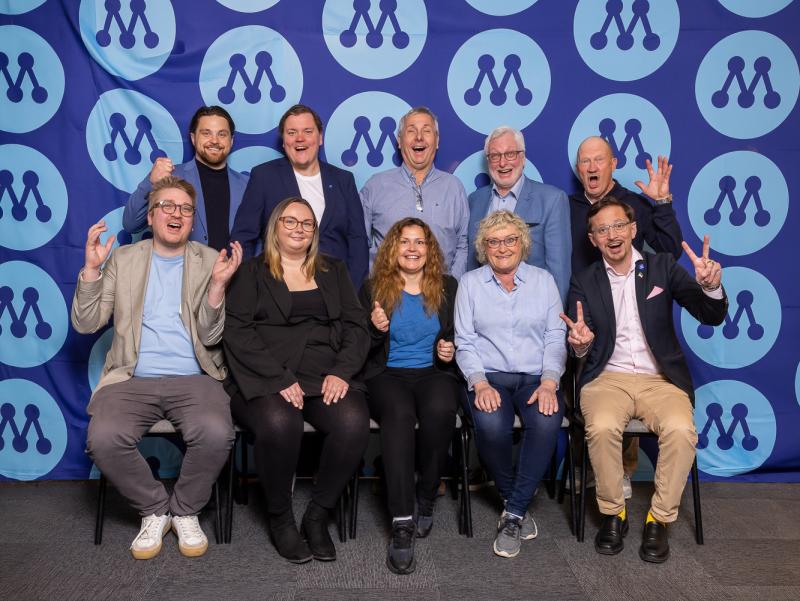 Tio personer poserar och ler i två rader framför en blå bakgrund med upprepade cirkelformade logotyper som föreställer ett stiliserat ”M”.