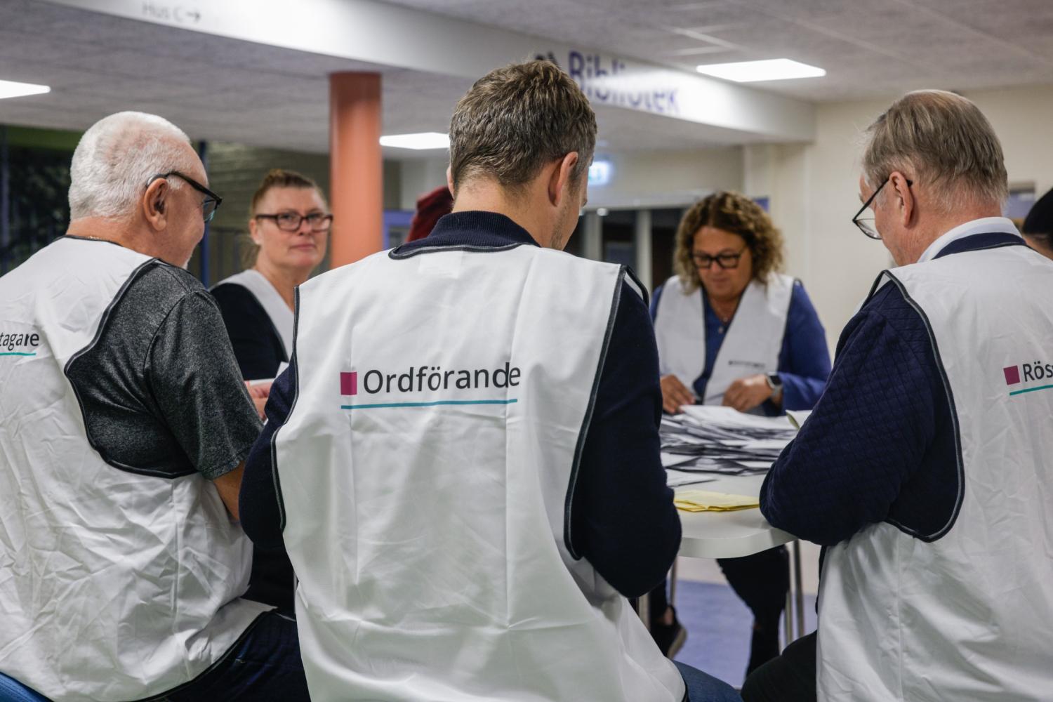 Fem personer i vita västar sitter vid ett bord och sorterar papper inomhus; de är troligen sysselsatta med rösträkning i samband med ett val eller med administrativt arbete.