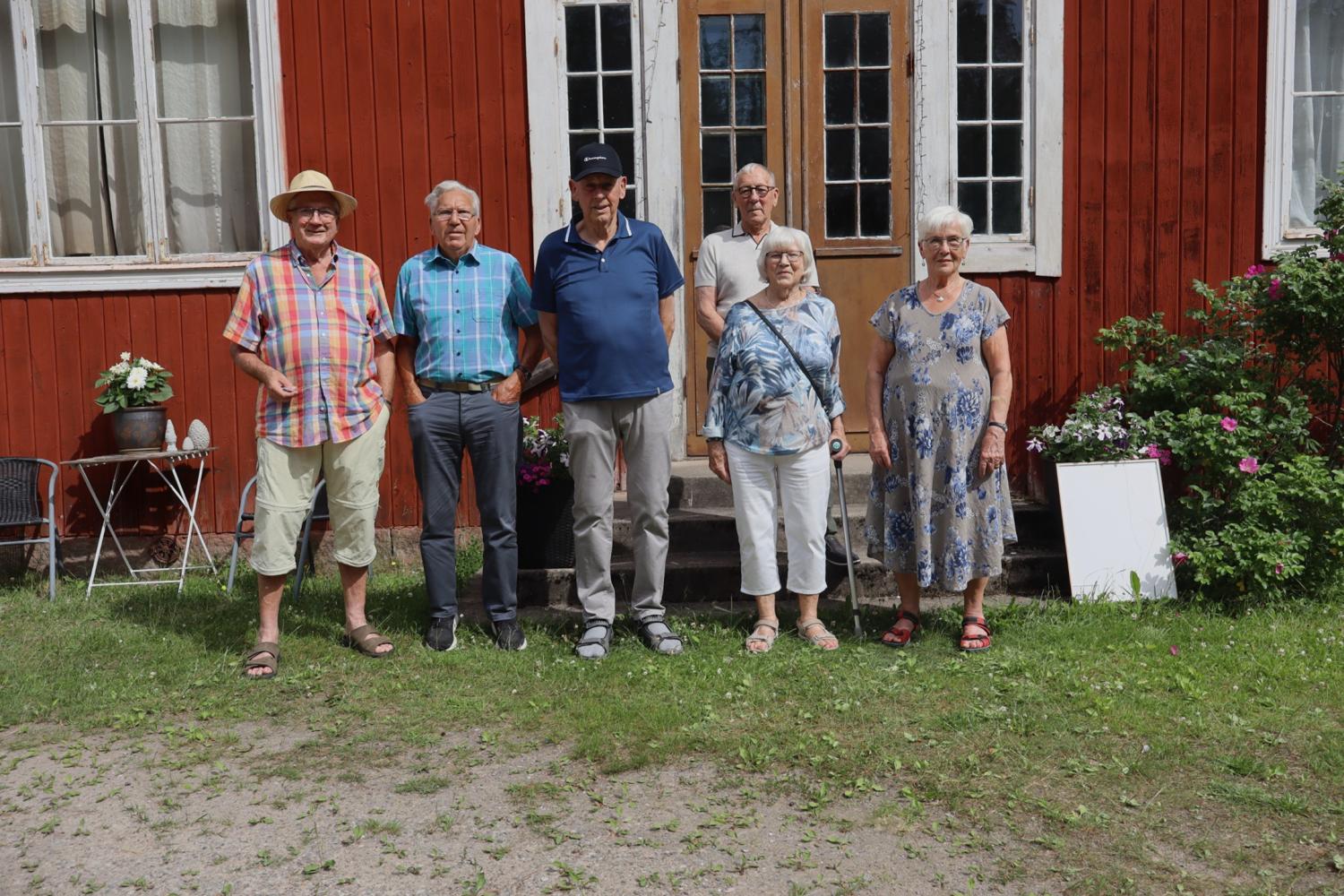 Sex äldre personer står på rad utanför en röd träbyggnad med stora fönster och en dubbeldörr, omgiven av grönt gräs och blommor.