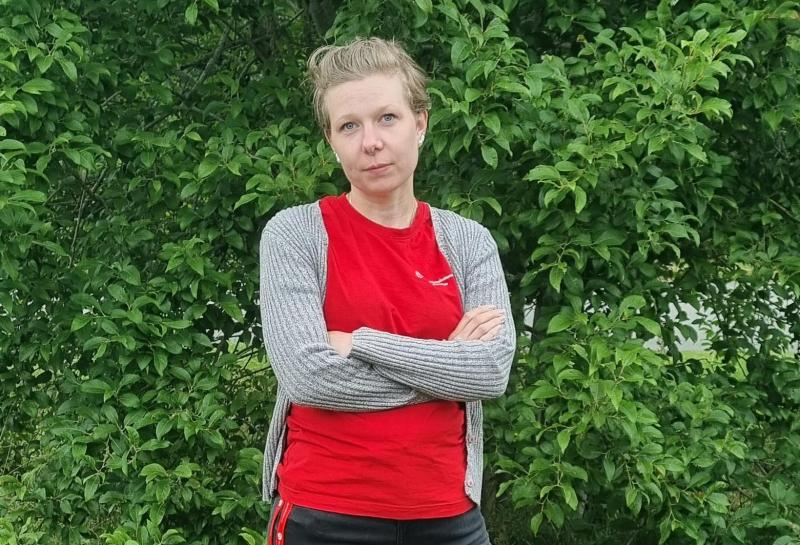 En kvinna med blont hår uppsatt i en knut, klädd i en röd tröja och en grå kofta, står med armarna i kors framför gröna, lövrika buskar.