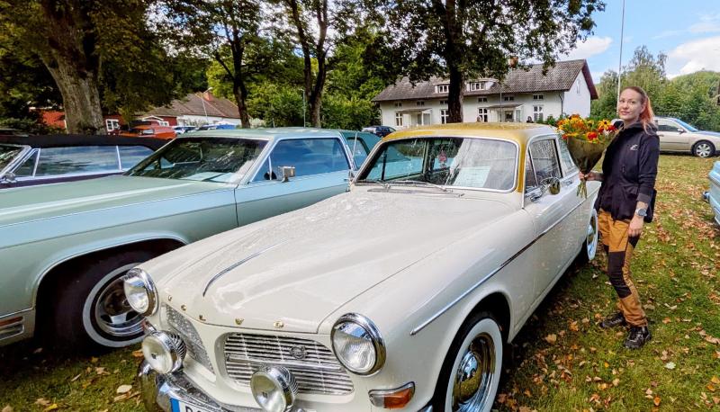 En kvinna står bredvid en vit Volvo av äldre modell som står parkerad på gräset, med andra klassiska bilar och träd i bakgrunden.