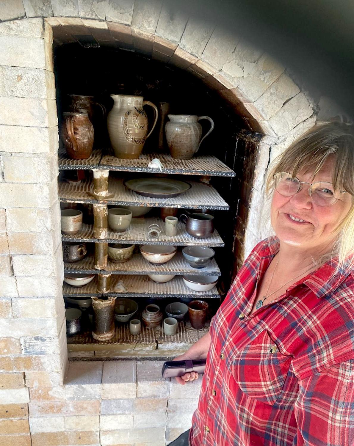 En kvinna i en rutig skjorta ler bredvid en ugn fylld med olika keramiska keramikbitar.