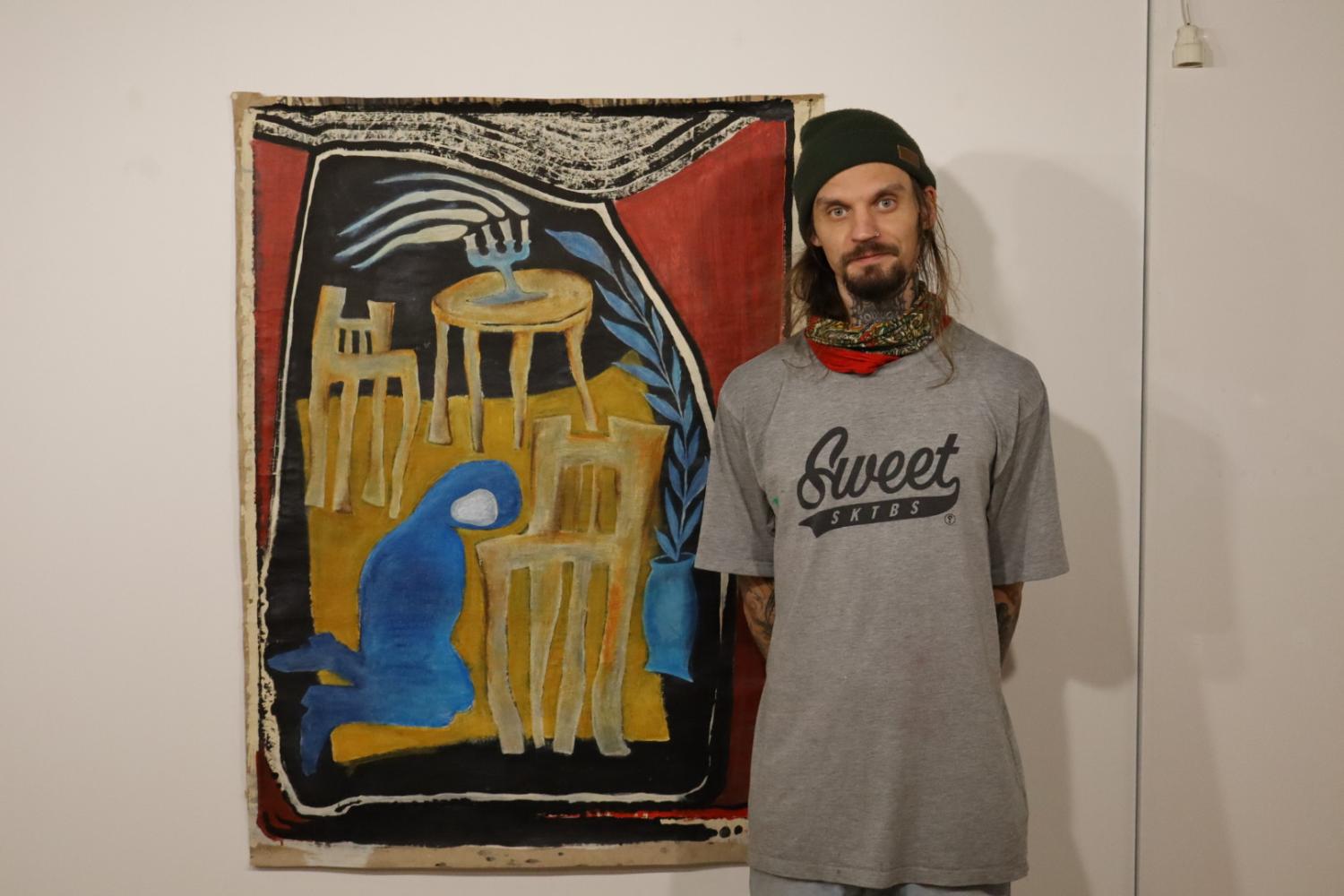 En person med långt hår står bredvid en abstrakt målning med stolar och en blå figur. De bär en grå t-shirt, mössa och röd halsduk.