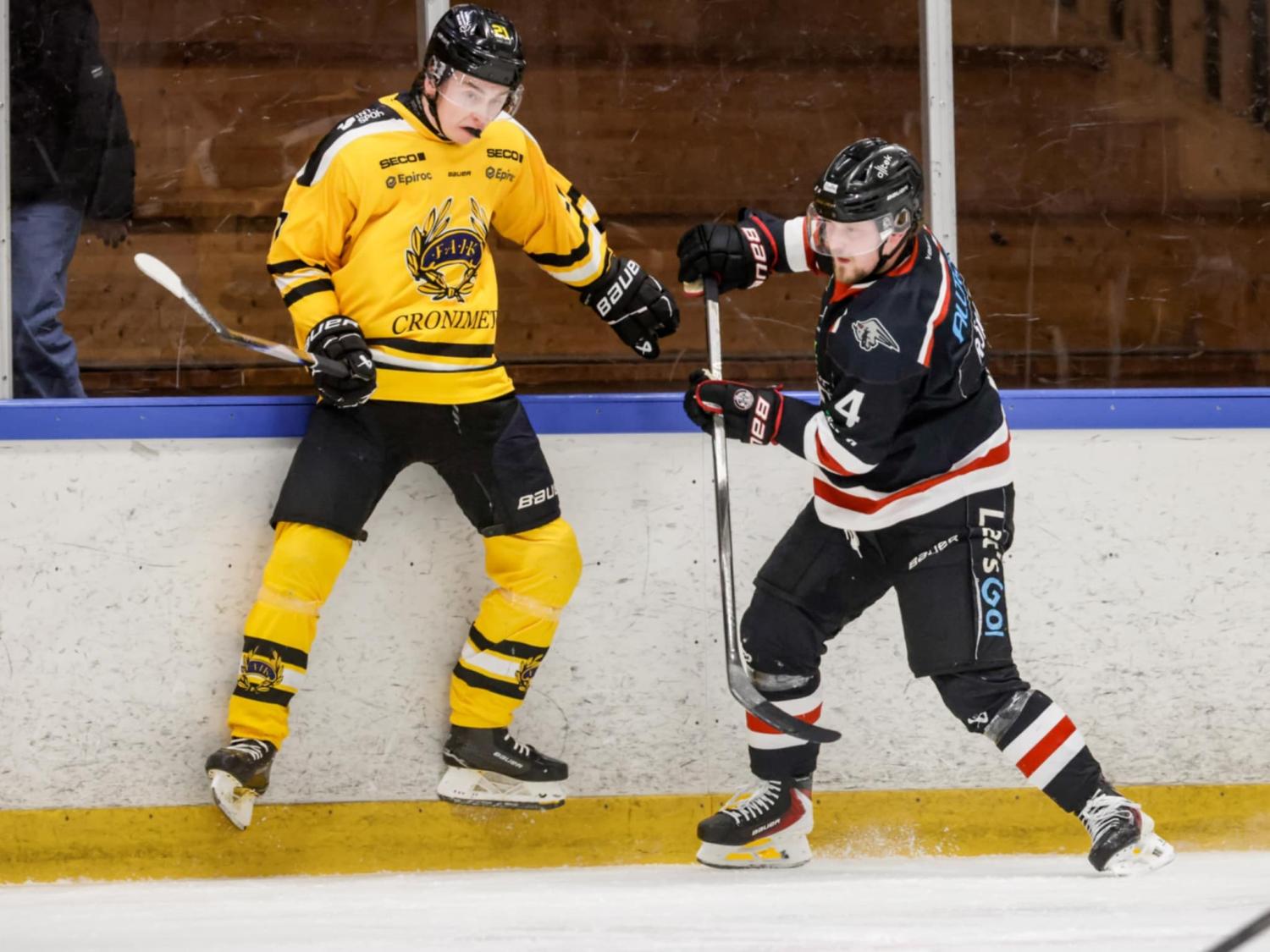 Två ishockeyspelare, en i gult och en i svart, tävlar om pucken nära sargen under en match.
