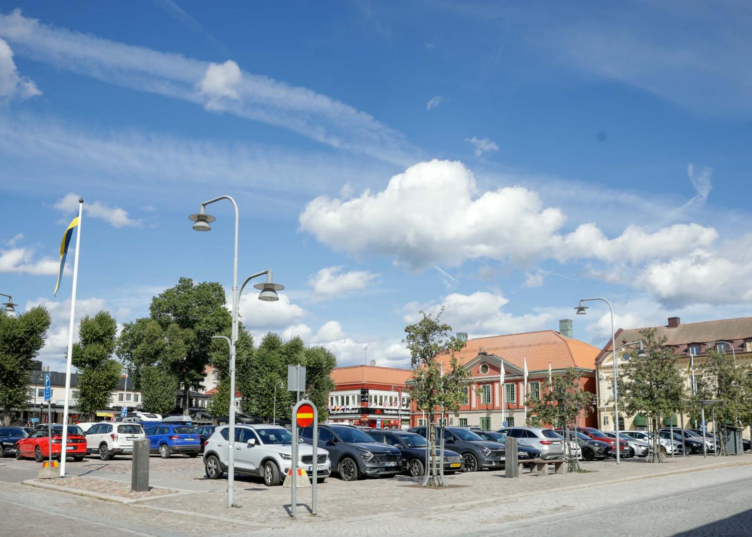 En parkeringsplats full av bilar en solig dag, med byggnader och träd i bakgrunden under en blå himmel med moln.