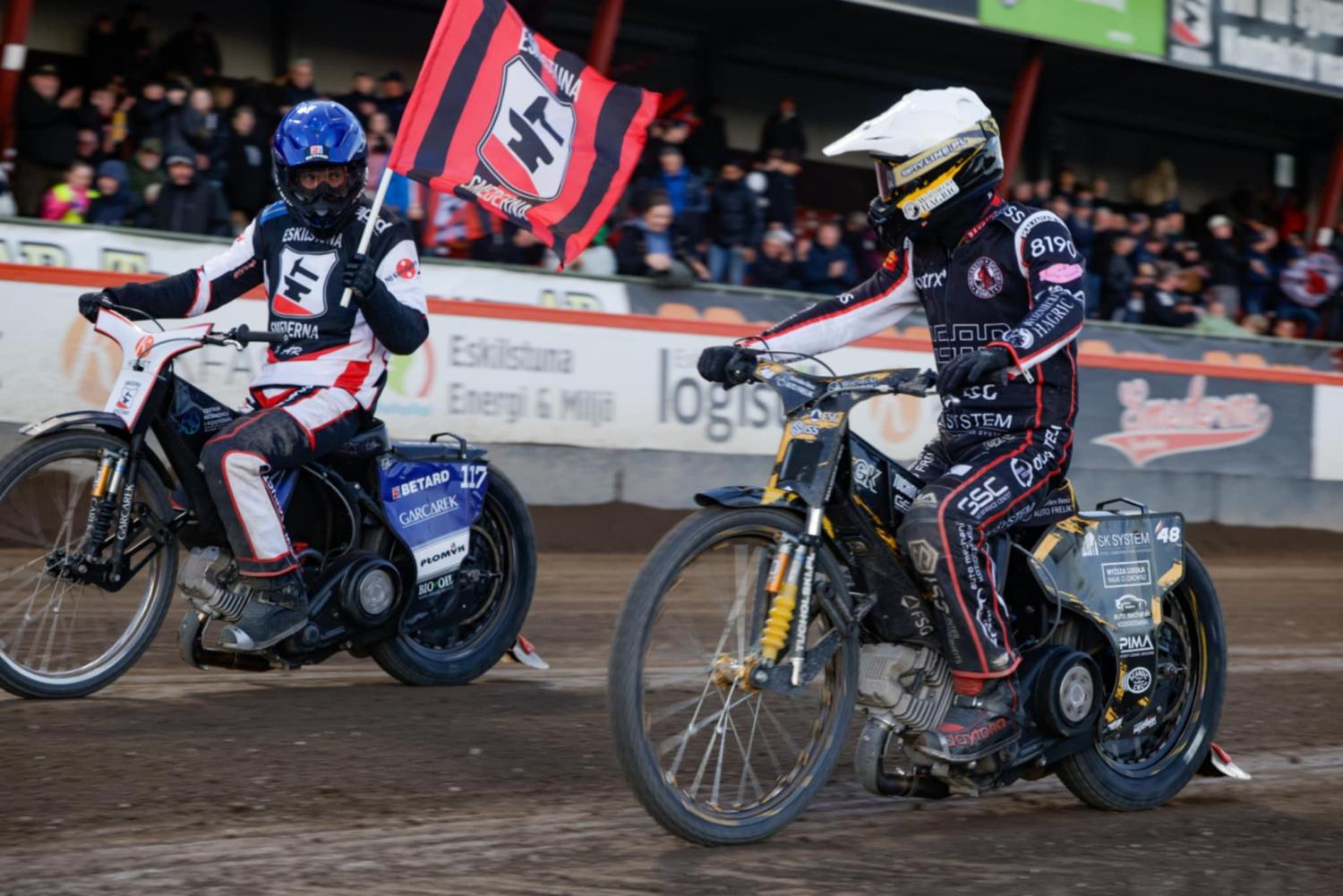 Två speedwayförare i tävlingskläder tävlar på en grusbana; den ena föraren håller i en röd och svart flagga. Åskådare tittar på från läktaren i bakgrunden.