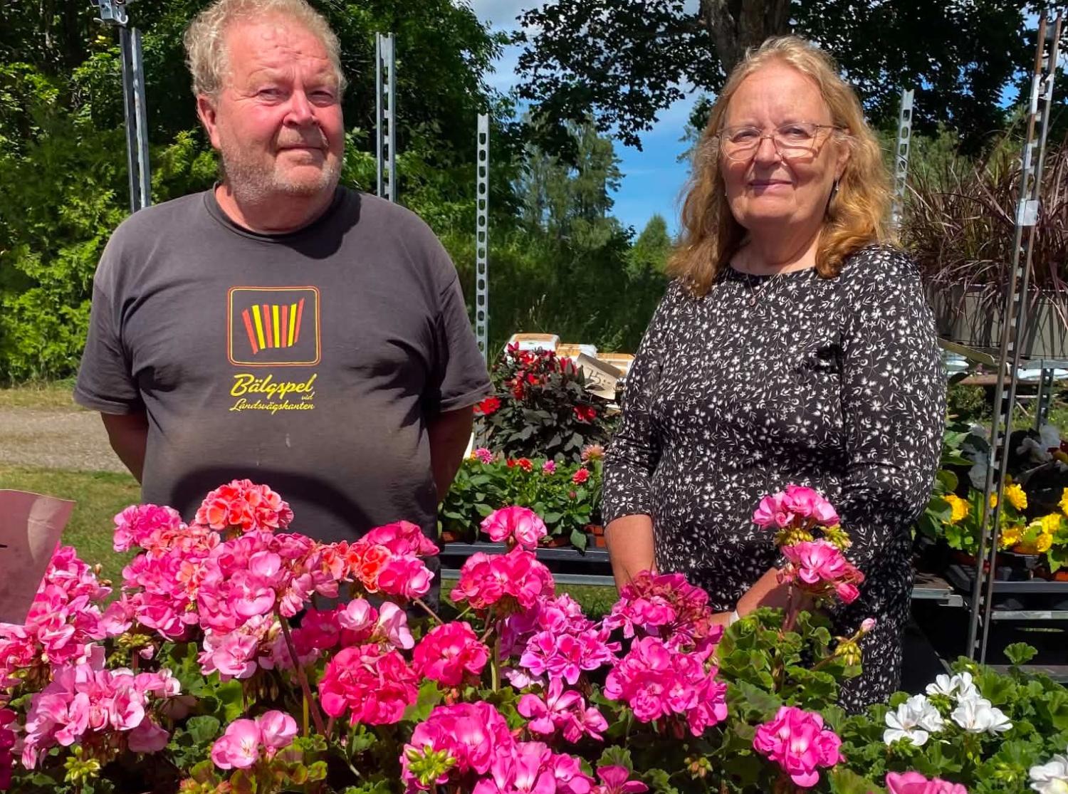 Två personer som står utomhus bakom ett bord med rosa och röda blommor, med grönska och krukväxter i bakgrunden.