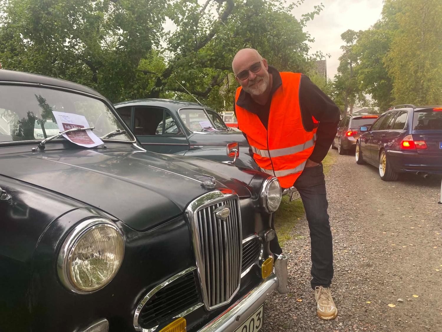 En man klädd i en knallorange säkerhetsväst står bredvid en klassisk svart bil på en grusväg kantad av andra fordon och träd.