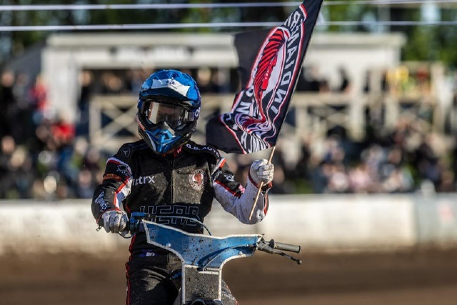 En speedwayförare i tävlingskläder och hjälm håller en lagflagga medan han kör motorcykel på en grusbana, med åskådare i bakgrunden.