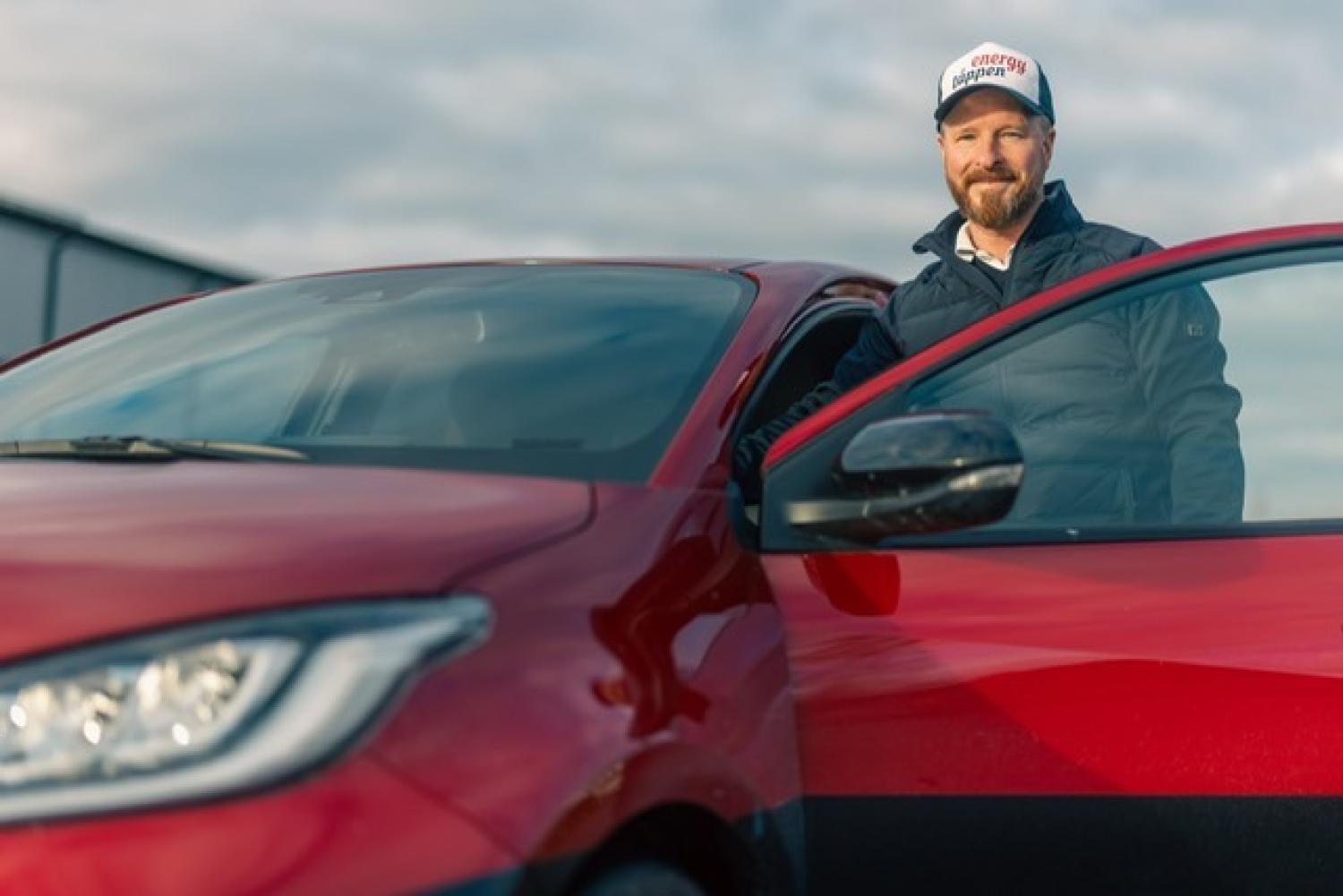 En man med keps och jacka står bredvid en öppen dörr på en röd bil och ler mot kameran under en molnig himmel.