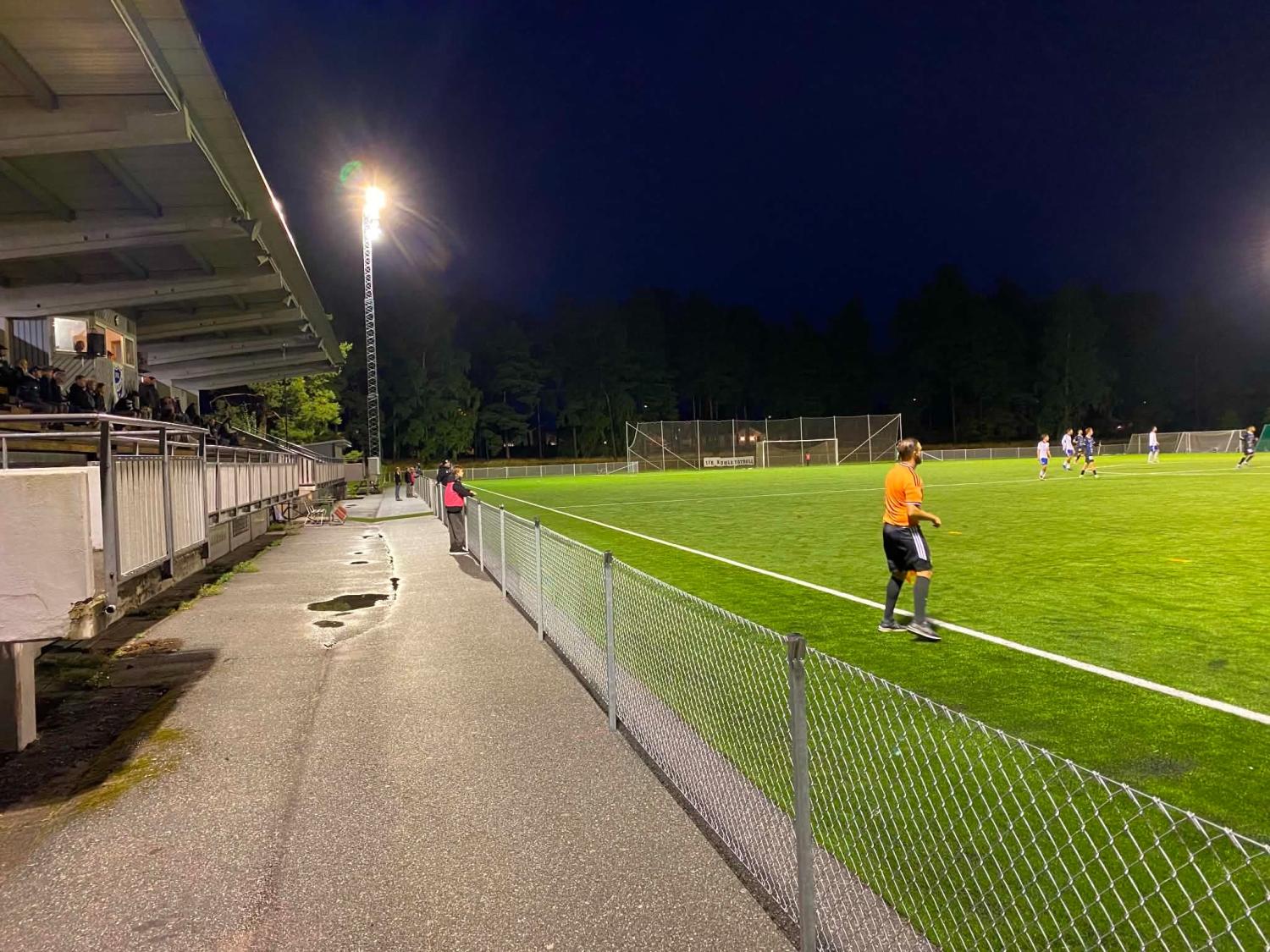 En fotbollsmatch spelas på kvällen på en upplyst plan, med en domare i orange och åskådare som sitter under en täckt läktare till vänster.