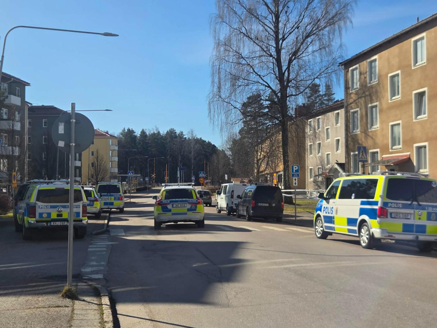 Flera svenska polisfordon står parkerade längs en bostadsgata i dagsljus, med några andra bilar och flerfamiljshus synliga.