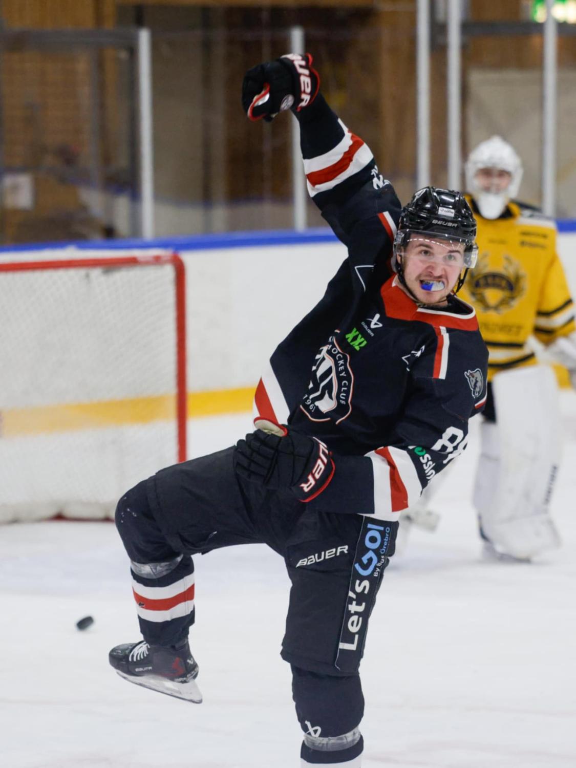 Hockeyspelare i svart uniform firar med uppsträckt arm på isen; motståndarmålvakten i gult står nära nätet i bakgrunden.