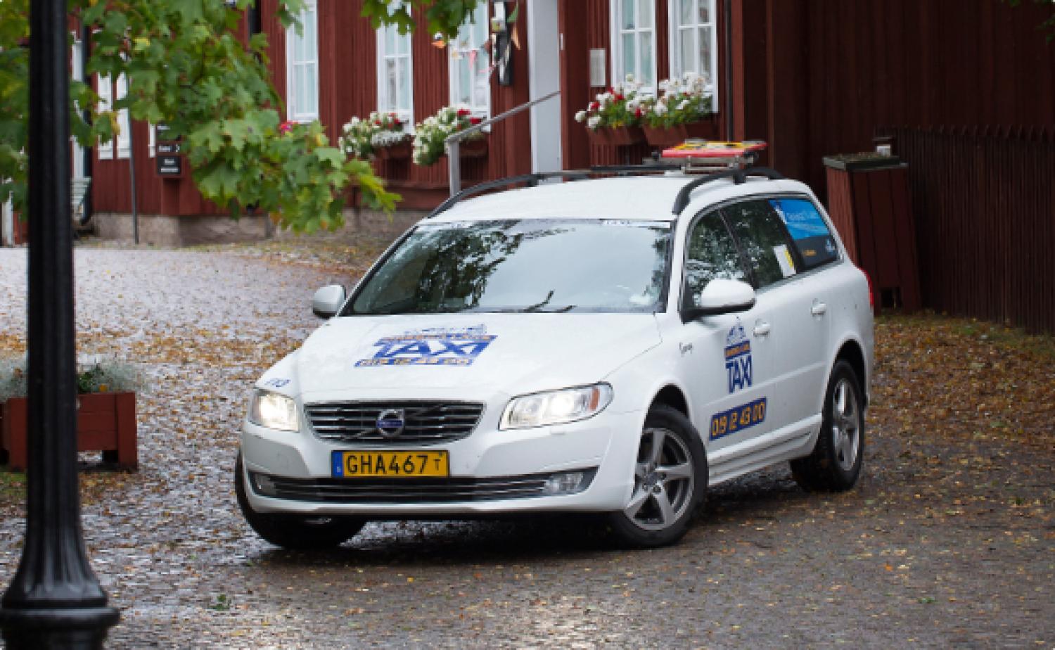 En vit Volvotaxi med takräcken och skyltar står parkerad på en kullerstensgata nära byggnader med röda fasader och grönt bladverk.