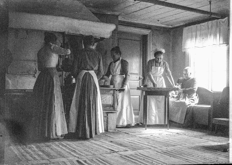 Fyra kvinnor står och arbetar i ett rustikt kök, medan en kvinna sitter vid ett bord vid fönstret i ett gammaldags rum med trägolv och synliga takbjälkar.