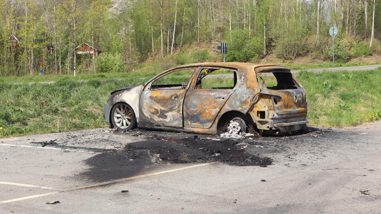 En svårt utbränd bil står övergiven på en parkeringsplats, omgiven av svartbrända bråte, med en gräsmatta och träd i bakgrunden.