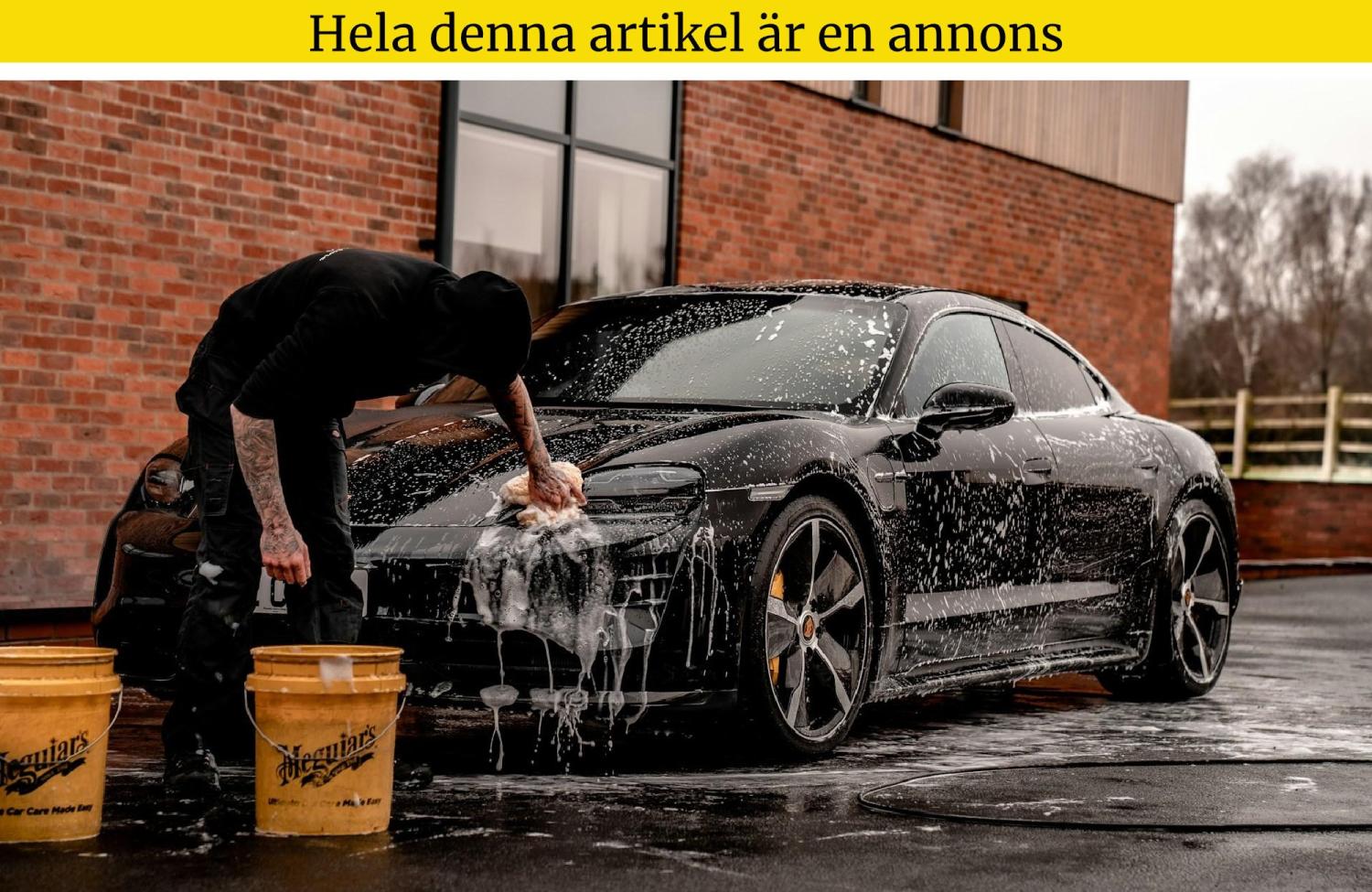 En person tvättar en svart sportbil med tvål och vatten utanför en tegelbyggnad med hjälp av två gula hinkar.
