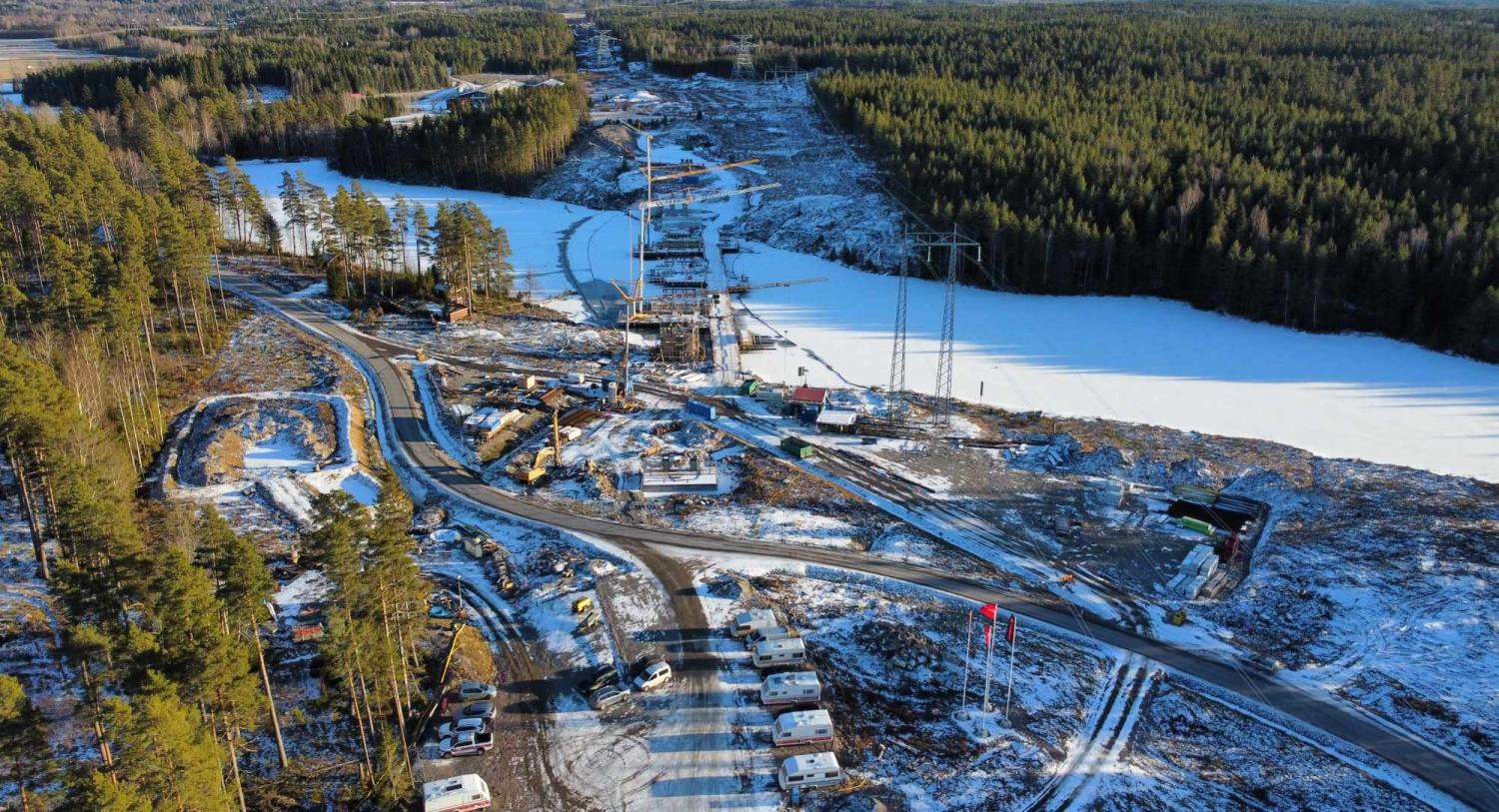 En snöig byggarbetsplats med maskiner och fordon omgiven av skogar. En flod rinner genom området.