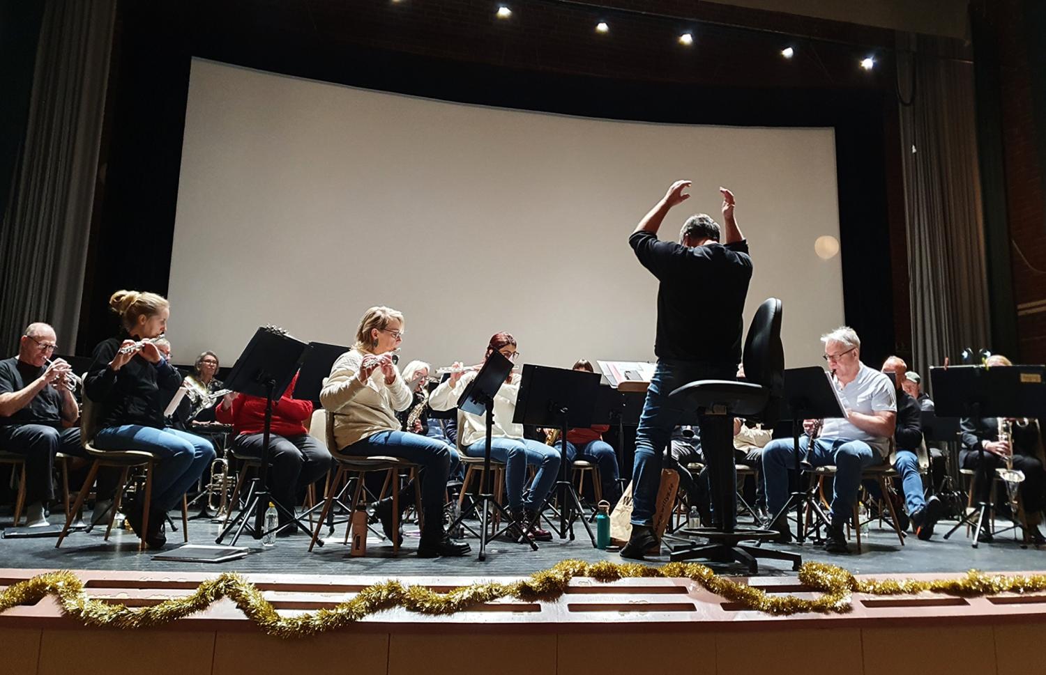 En dirigent leder en sittande orkester under en repetition på scenen; musiker spelar olika blåsinstrument med notställ framför sig.
