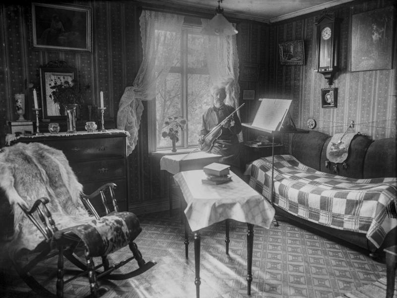 En äldre person sitter på en soffa och spelar fiol i ett gammaldags, vackert inrett rum med en gungstol, ett bord, ett piano och inramade tavlor på väggarna.