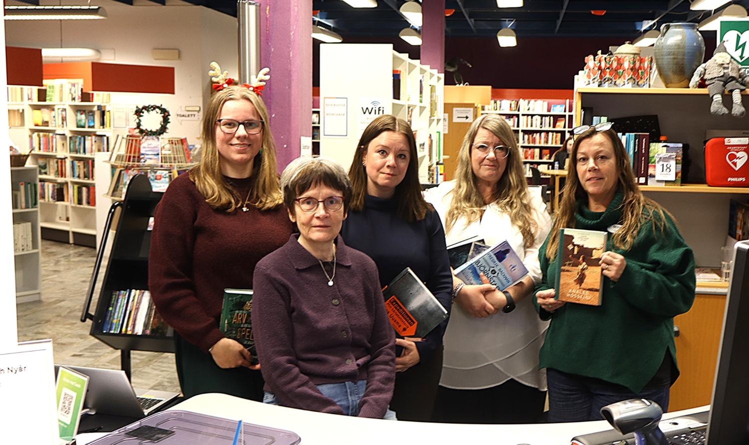 Fem kvinnor står tillsammans i ett bibliotek, var och en med en bok i handen, med bokhyllor och biblioteksutrustning i bakgrunden.
