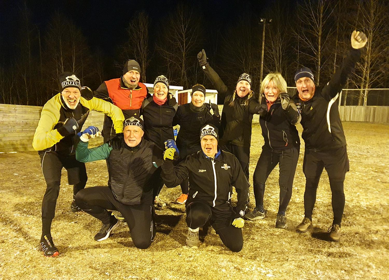 Nio vuxna personer klädda i sportiga vinterkläder poserar utomhus på natten på ett frostigt fält, ler och höjer armarna energiskt.